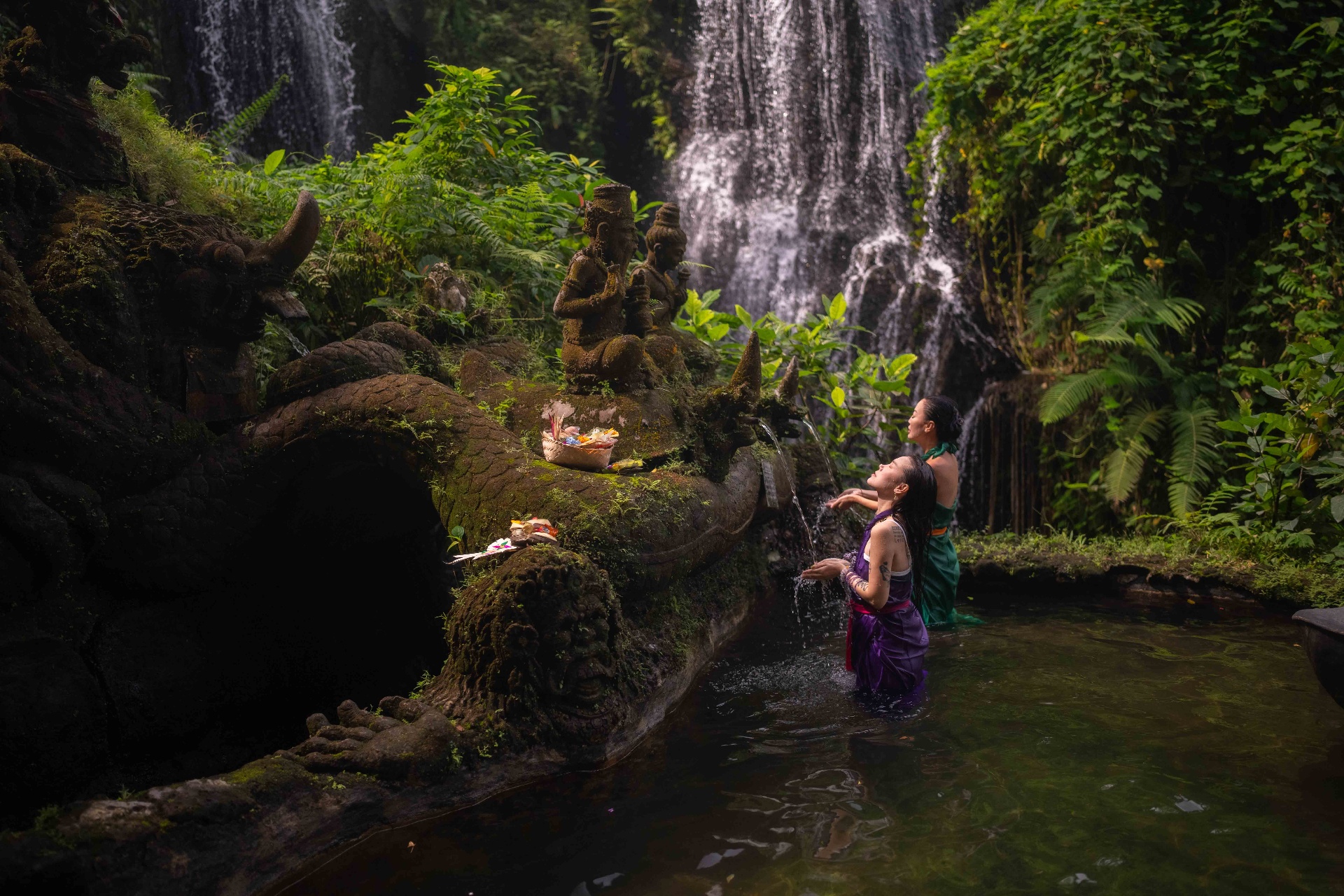 Women receive a sacred water blessing at a moss-carved shrine beneath a waterfall