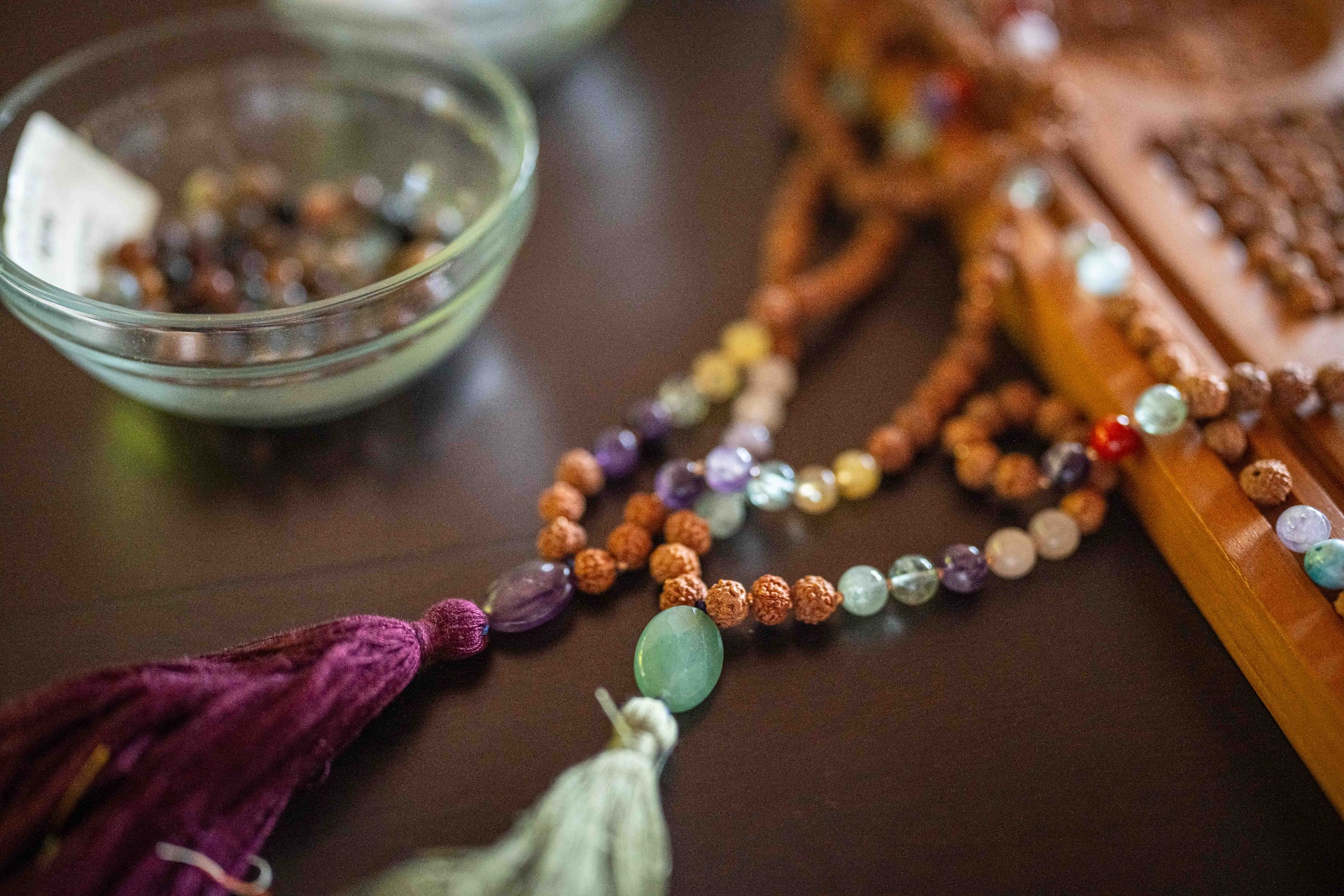 Close-up of a mala strand with coloured gemstone beads and silk tassels beside a bowl of offerings