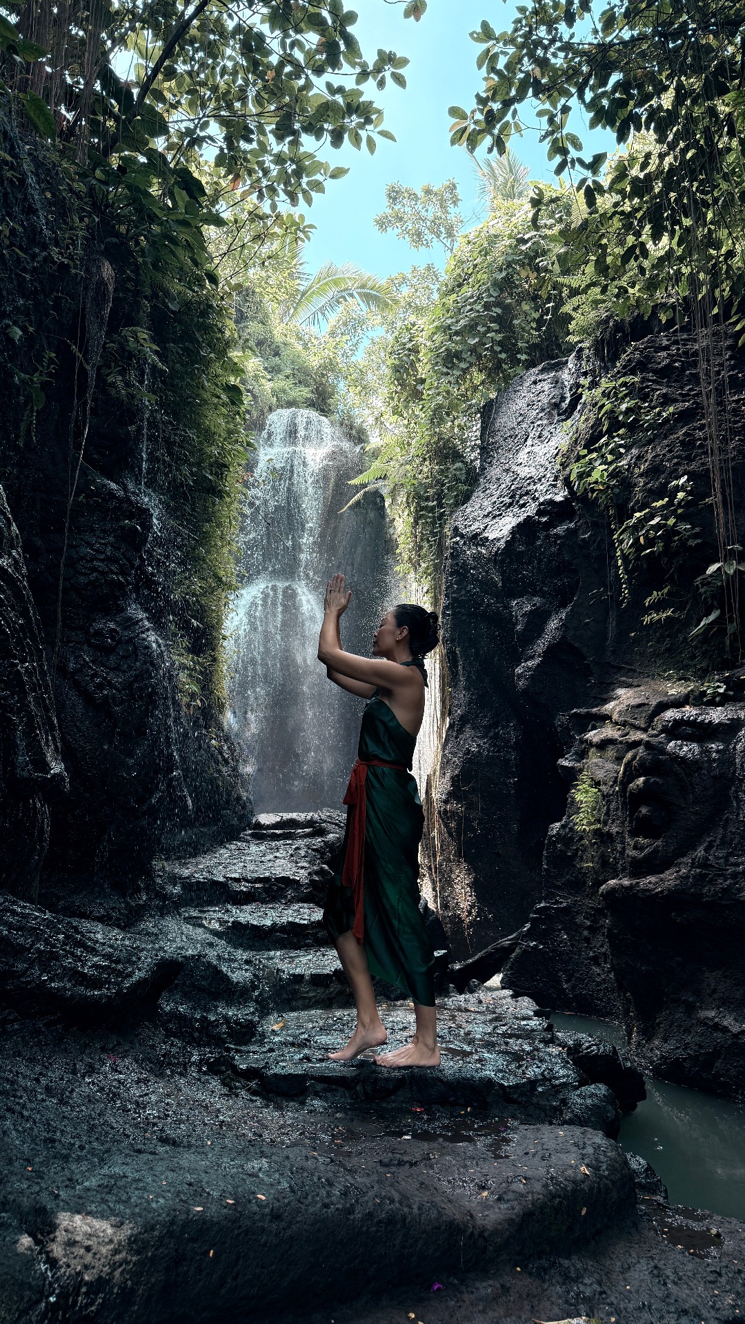 An outstretched hand beneath flowing holy water with canang sari flower offerings on a moss-covered stone shrine